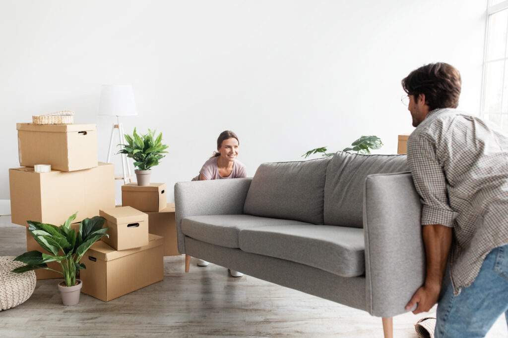 Buy To Let Mortgages Smiling millennial caucasian couple in casual carry sofa in room with packed cardboard boxes, planning interior